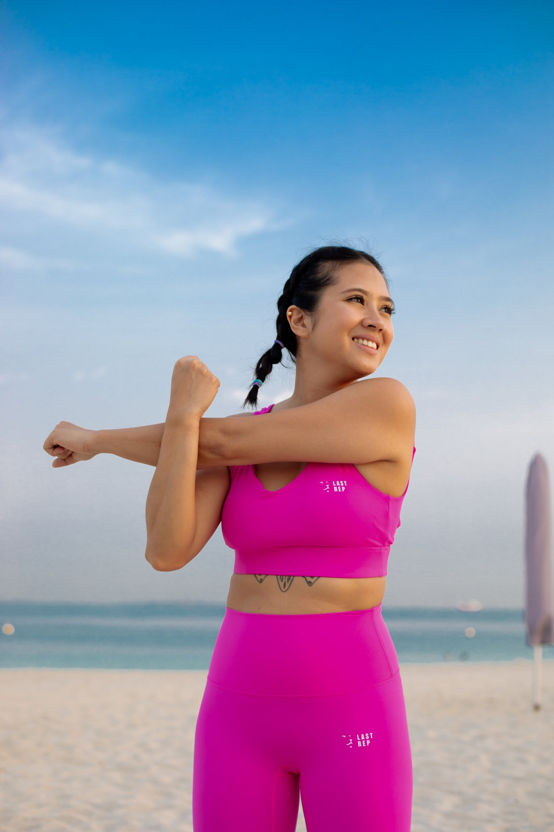 Woman in pink athletic wear stretching on a beach with a clear blue sky.